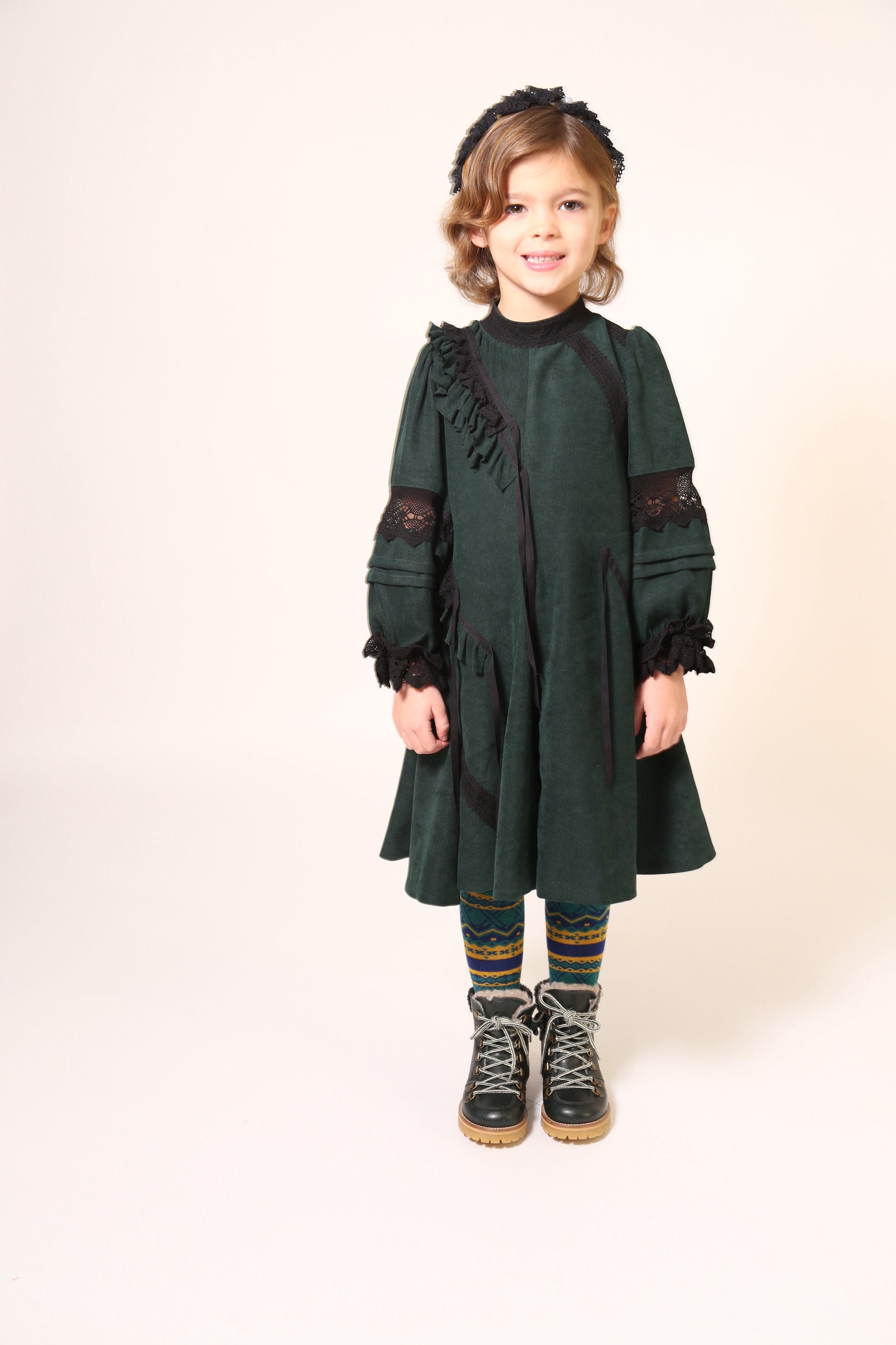 Girl wearing a party dress in soft Liquid Canvas fabric with diagonal ruffle details. Lace trim and ribbon at ruffles, neckline, elbows, and cuffs. Mock collar with button closure and invisible zipper at back. Big A-flare sweep at godet panels, cinched long sleeves with fold-up details. Colour: Celtic,Tia Cibani.