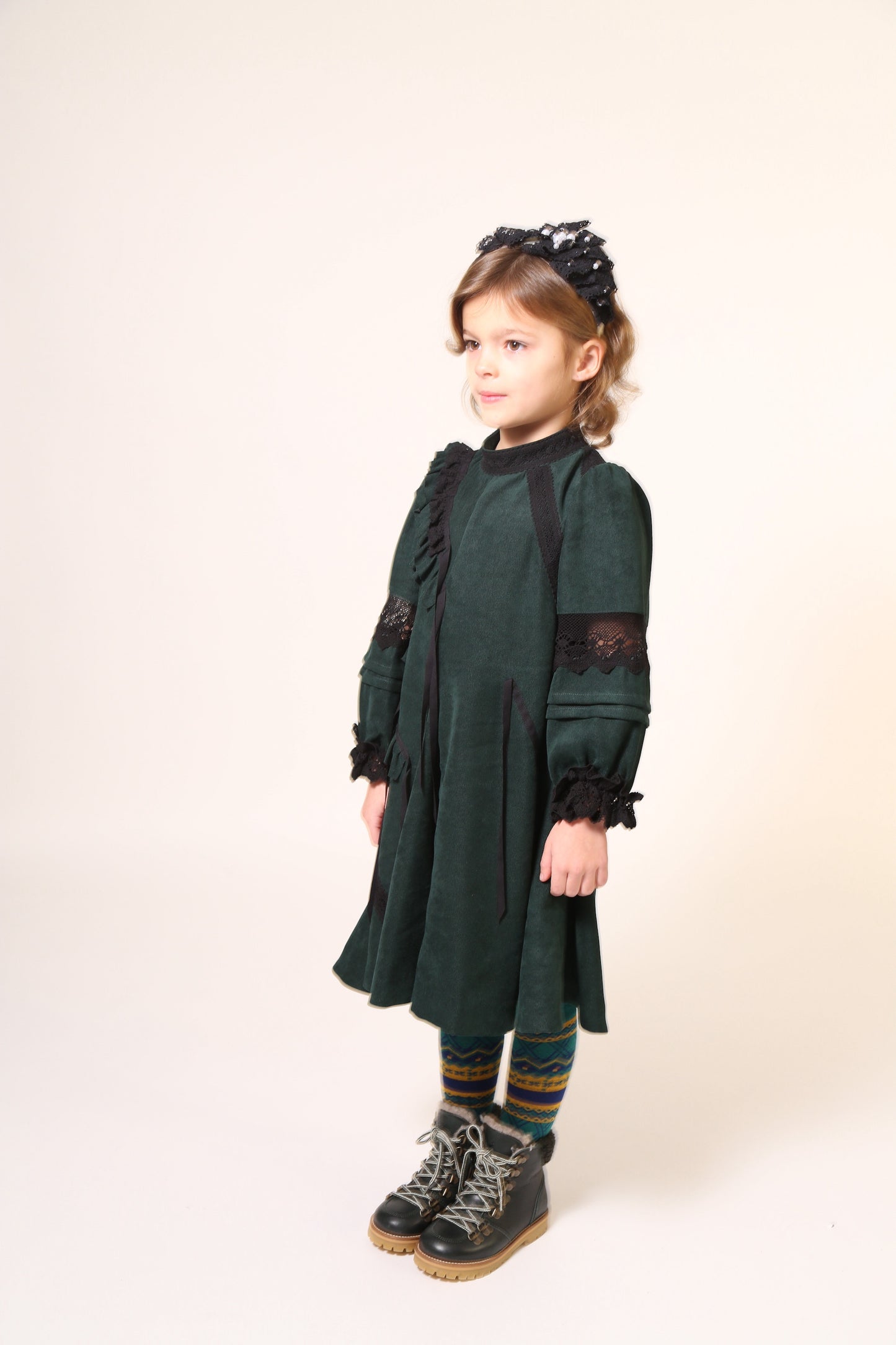 Girl wearing a party dress in soft Liquid Canvas fabric with diagonal ruffle details. Lace trim and ribbon at ruffles, neckline, elbows, and cuffs. Mock collar with button closure and invisible zipper at back. Big A-flare sweep at godet panels, cinched long sleeves with fold-up details. Colour: Celtic,Tia Cibani.