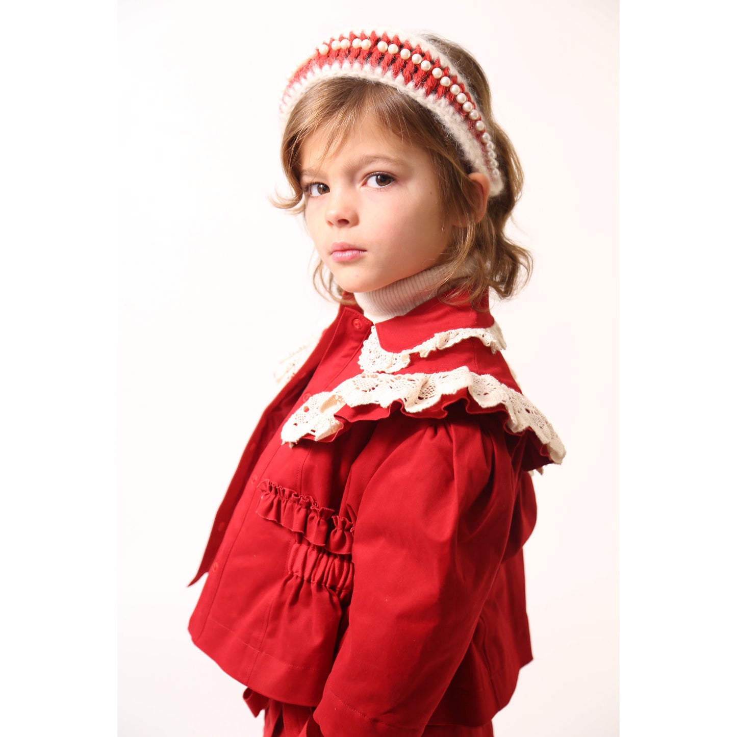 Girl wearing a white and rouge oversized headband wrapped and crocheted with mohair and wool yarn, adorned with pearls at the crown, Tia Cibani. / Girl wearing a charming volume topper jacket in soft Chamois Cotton Twill with contrasting lace embellishments, Red and White, Tia Cibani.