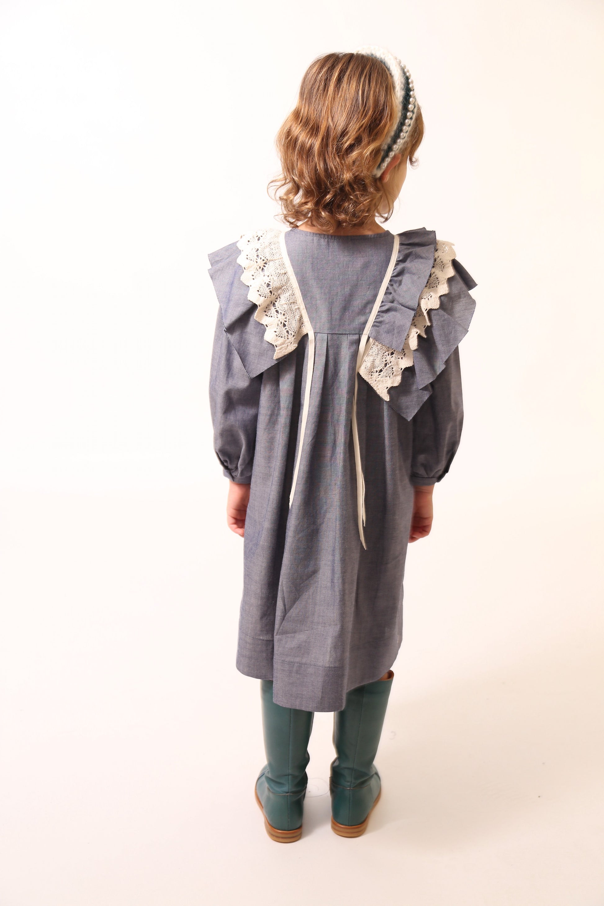 Girl wearing a voluminous dress in unlined Chambray Cotton Shirting with 3-D self ruffles at shoulders and delicate lace ruffles overlay. Decorative ribbons from ruffles. Long cinched sleeves, tealength silhouette, henley snap placket at center front neckline. Colour: Blue, Tia Cibani.