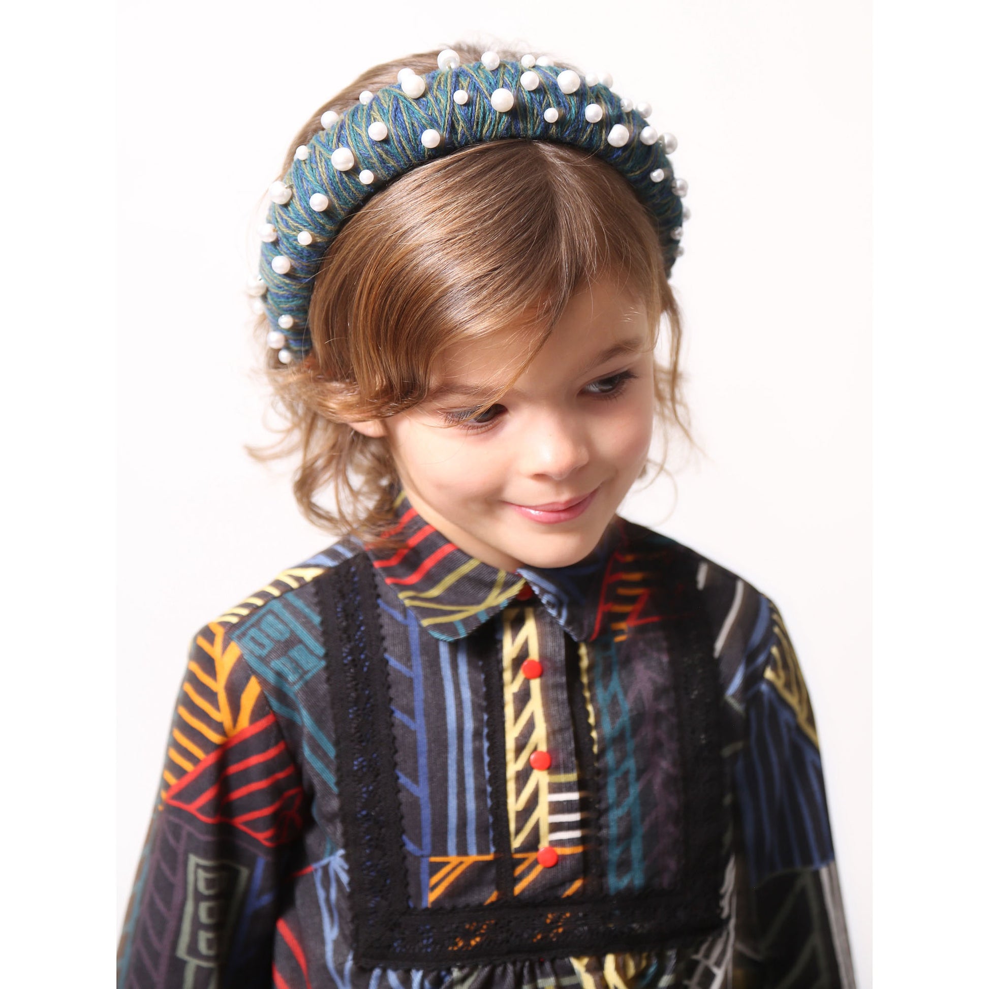 Child wearing a headband covered in blue and green thread with various pearls attached, by Tia Cibani.