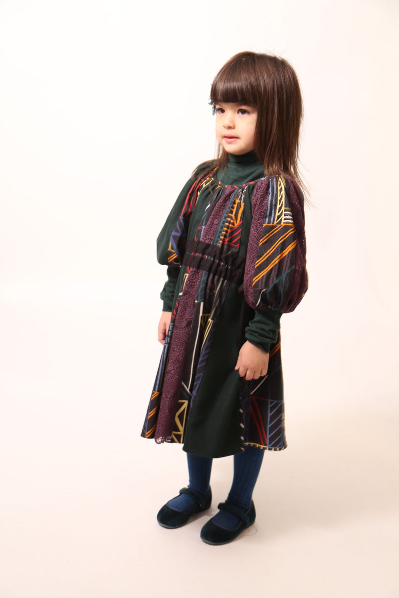 Girl wearing Patchworked frock for girls with signature print, embroidered lace, and liquid canvas fabric. Soft cotton lining, layered Argyle Tulle for volume, cinched sleeves, scoop neckline, and adjustable ribbon waist detail. Perfect for special occasions. Dark Rainbow,Tia Cibani.