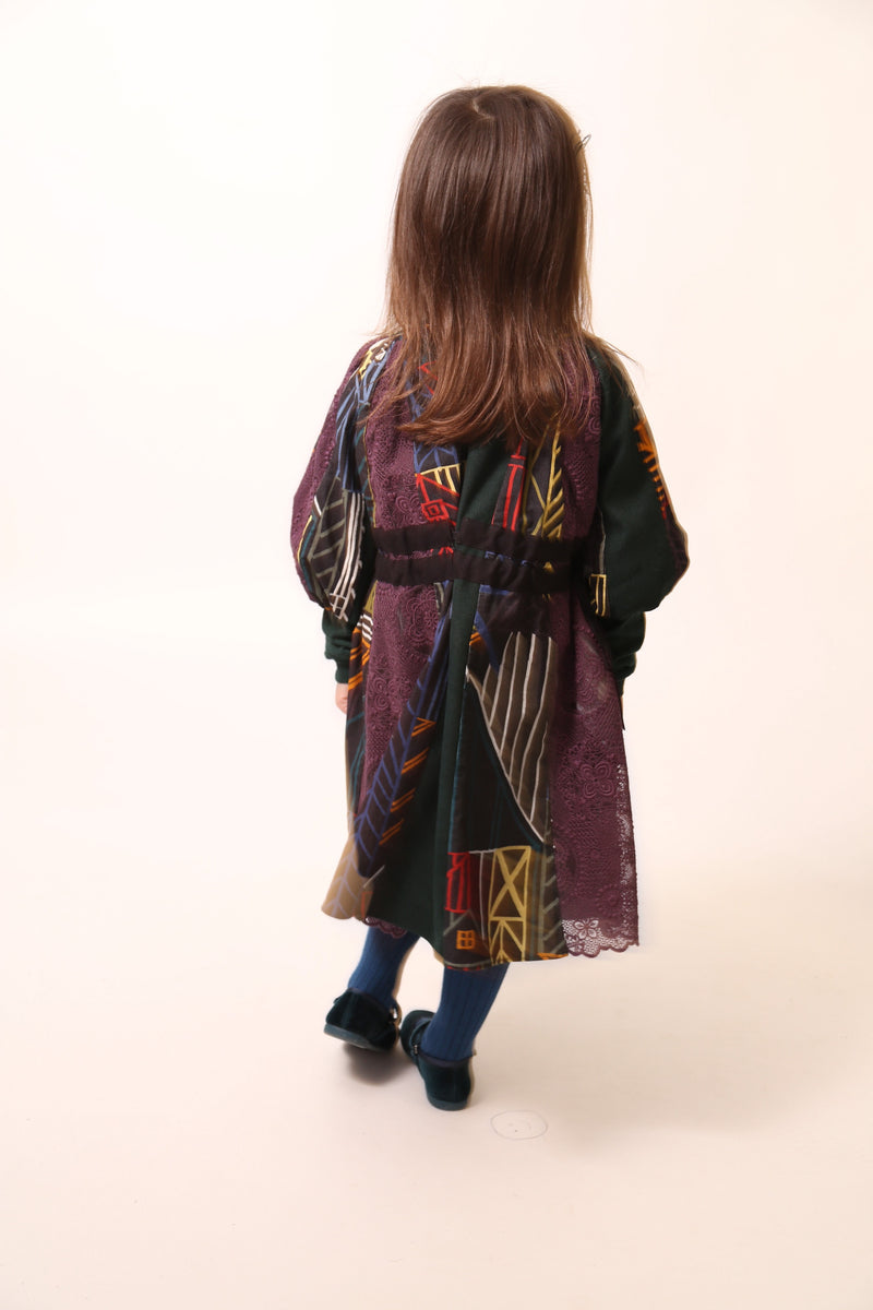 Girl wearing Patchworked frock for girls with signature print, embroidered lace, and liquid canvas fabric. Soft cotton lining, layered Argyle Tulle for volume, cinched sleeves, scoop neckline, and adjustable ribbon waist detail. Perfect for special occasions. Dark Rainbow,Tia Cibani.