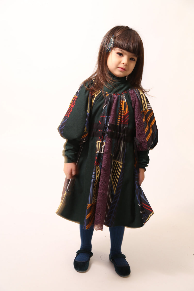 Girl wearing Patchworked frock for girls with signature print, embroidered lace, and liquid canvas fabric. Soft cotton lining, layered Argyle Tulle for volume, cinched sleeves, scoop neckline, and adjustable ribbon waist detail. Perfect for special occasions. Dark Rainbow,Tia Cibani.