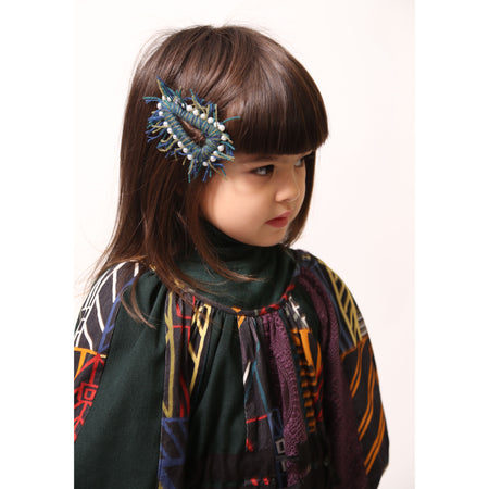 Girl wearing a oversized hair clip with a sturdy metal snap clip base, wrapped and fringed in colorful blue and green wool yarn, decorated with pearls for a fun finish, by Tia Cibani.