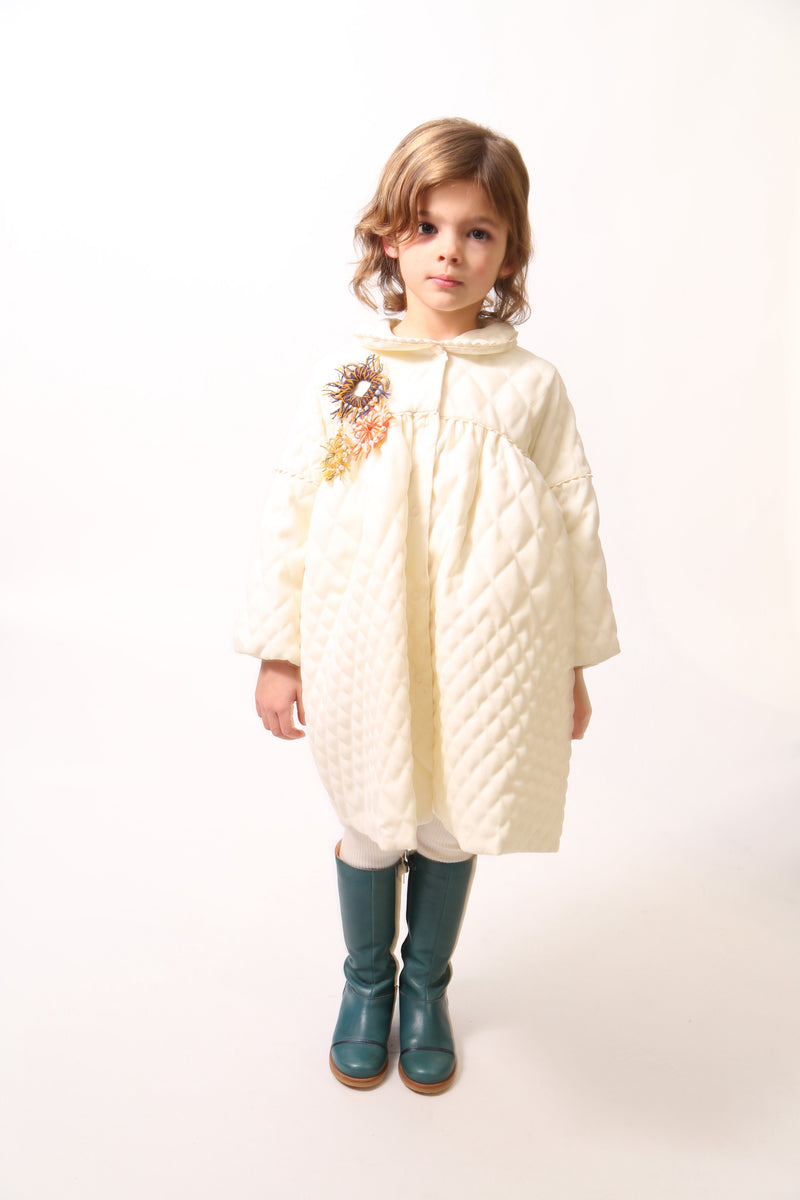 Girl wearing a Peter Pan collar tealength dress with long cinched sleeves, diamond harlequin pattern tufting, warm padding, and snap button closure. Ric rac ribbon trimming adds charm. Can double as an overcoat. Colour: White, Tia Cibani.