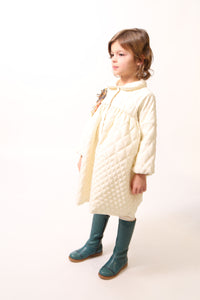 Girl wearing a Peter Pan collar tealength dress with long cinched sleeves, diamond harlequin pattern tufting, warm padding, and snap button closure. Ric rac ribbon trimming adds charm. Can double as an overcoat. Colour: White, Tia Cibani.