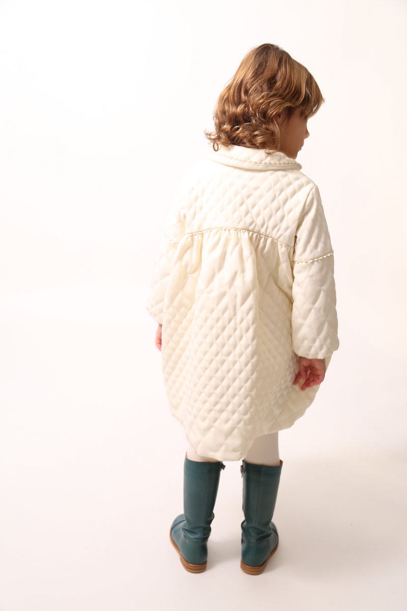 Girl wearing a Peter Pan collar tealength dress with long cinched sleeves, diamond harlequin pattern tufting, warm padding, and snap button closure. Ric rac ribbon trimming adds charm. Can double as an overcoat. Colour: White, Tia Cibani.