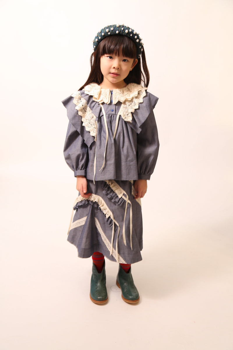 A girl wearing an everyday blouse in soft Chambray Cotton Shirting with 3-D self ruffles at shoulder, lace overlay, and lace trim at neckline for two-tiered collar. Airy, lightweight fit with henley snap placket at center front. Cinched long sleeves, minimal ribbons on ruffles. Colour: Blue, Tia Cibani.