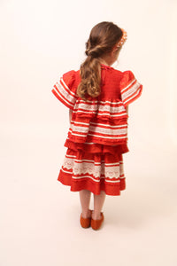 Girl wearing a top in luxurious Taffeta with elasticized smocking at neckline, ruffle detail, and embroidered lace trim and ribbon at body and flutter sleeves. Tonal ribbon ties at the back of the neck. Unlined for lightweight feel. Colour: Rouge and white, Tia Cibani. / A girl wearing Tealength two-tiered taffeta skirt with elasticized smocking at the waistband, embroidered lace trim, and ribbon at each tier. Unlined for lightweight feel. Colour: Rouge and White, Tia Cibani.