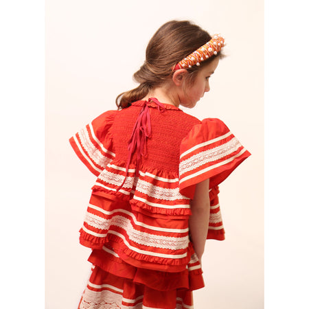 Girl wearing a top in luxurious Taffeta with elasticized smocking at neckline, ruffle detail, and embroidered lace trim and ribbon at body and flutter sleeves. Tonal ribbon ties at the back of the neck. Unlined for lightweight feel. Colour: Rouge and white, Tia Cibani.
