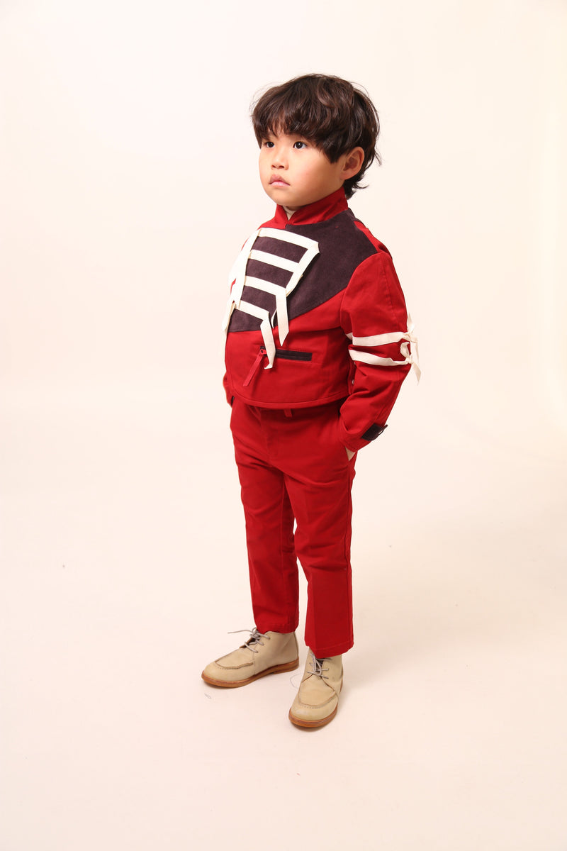 Boy wearing a bold and stylish Malok Military Jacket for boys, inspired by traditional military dress. Patchworked with cotton twill and velvet accents. Features zip welt pockets, yoke at back, snap tabs at sleeves, and double-breasted snap closure for comfort and charm. Relaxed box fit for easy layering. Textured structured ribbon details on front and sleeves with free-hanging tassels for contemporary flair. Scoop hem, stand-up collar. Perfect with Malok Slim Fit Trousers. Rouge, Tia Cibani.