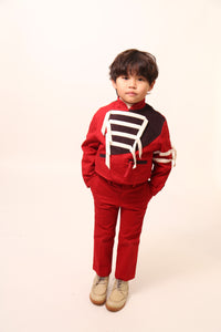 Boy wearing a bold and stylish Malok Military Jacket for boys, inspired by traditional military dress. Patchworked with cotton twill and velvet accents. Features zip welt pockets, yoke at back, snap tabs at sleeves, and double-breasted snap closure for comfort and charm. Relaxed box fit for easy layering. Textured structured ribbon details on front and sleeves with free-hanging tassels for contemporary flair. Scoop hem, stand-up collar. Perfect with Malok Slim Fit Trousers. Rouge, Tia Cibani.