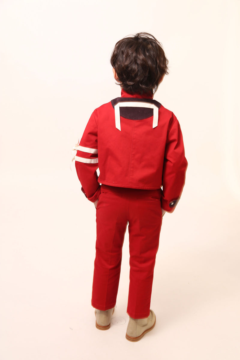 Boy wearing a bold and stylish Malok Military Jacket for boys, inspired by traditional military dress. Patchworked with cotton twill and velvet accents. Features zip welt pockets, yoke at back, snap tabs at sleeves, and double-breasted snap closure for comfort and charm. Relaxed box fit for easy layering. Textured structured ribbon details on front and sleeves with free-hanging tassels for contemporary flair. Scoop hem, stand-up collar. Perfect with Malok Slim Fit Trousers. Rouge, Tia Cibani.