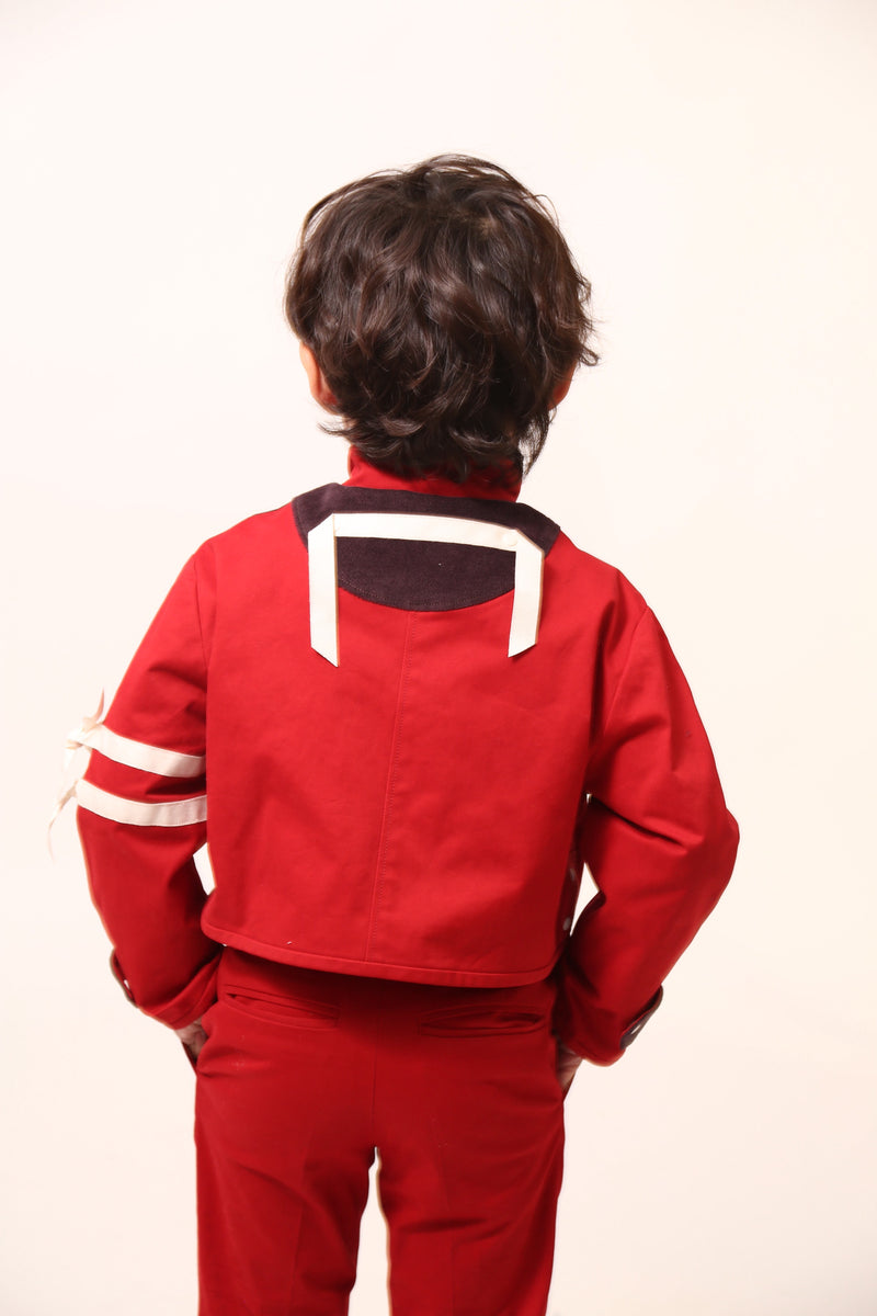 Boy wearing a bold and stylish Malok Military Jacket for boys, inspired by traditional military dress. Patchworked with cotton twill and velvet accents. Features zip welt pockets, yoke at back, snap tabs at sleeves, and double-breasted snap closure for comfort and charm. Relaxed box fit for easy layering. Textured structured ribbon details on front and sleeves with free-hanging tassels for contemporary flair. Scoop hem, stand-up collar. Perfect with Malok Slim Fit Trousers. Rouge, Tia Cibani.