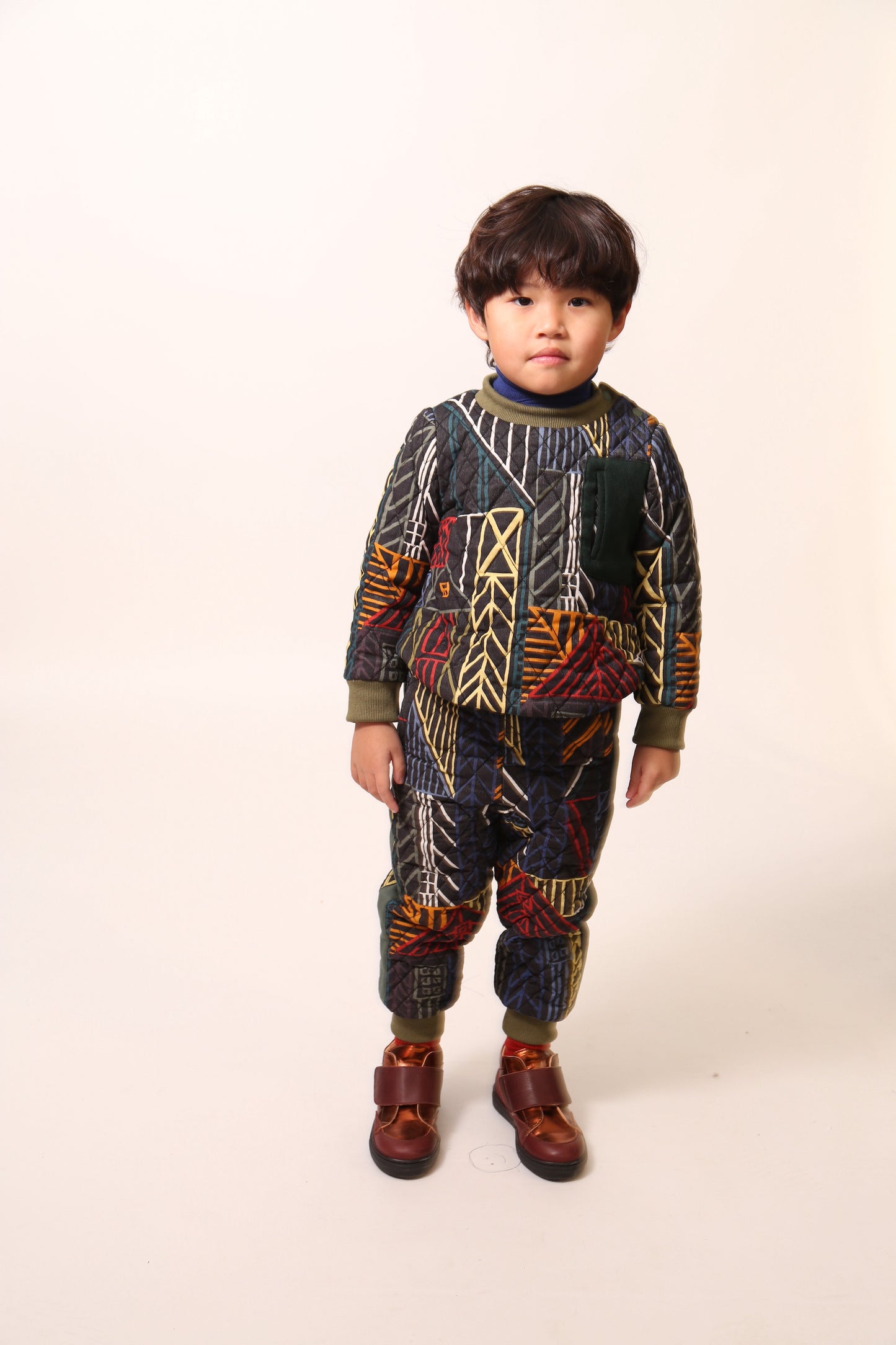 Boy wearing sporty track pants for boys in Normanz print, with harlequin diamond tufting and heavy padding for warmth. Pull-on elastic ribbed waistband, side seam pockets, ribbed leg openings for a snug fit. Contrast patch pocket at back, ribbons lining sides. Pair with Harlequin Padded Zip Sweatshirt for full rainbow track suit look. Dark rainbow, Tia Cibani.