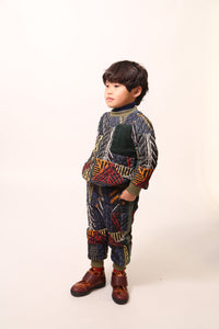 Boy wearing sporty track pants for boys in Normanz print, with harlequin diamond tufting and heavy padding for warmth. Pull-on elastic ribbed waistband, side seam pockets, ribbed leg openings for a snug fit. Contrast patch pocket at back, ribbons lining sides. Pair with Harlequin Padded Zip Sweatshirt for full rainbow track suit look. Dark rainbow, Tia Cibani.