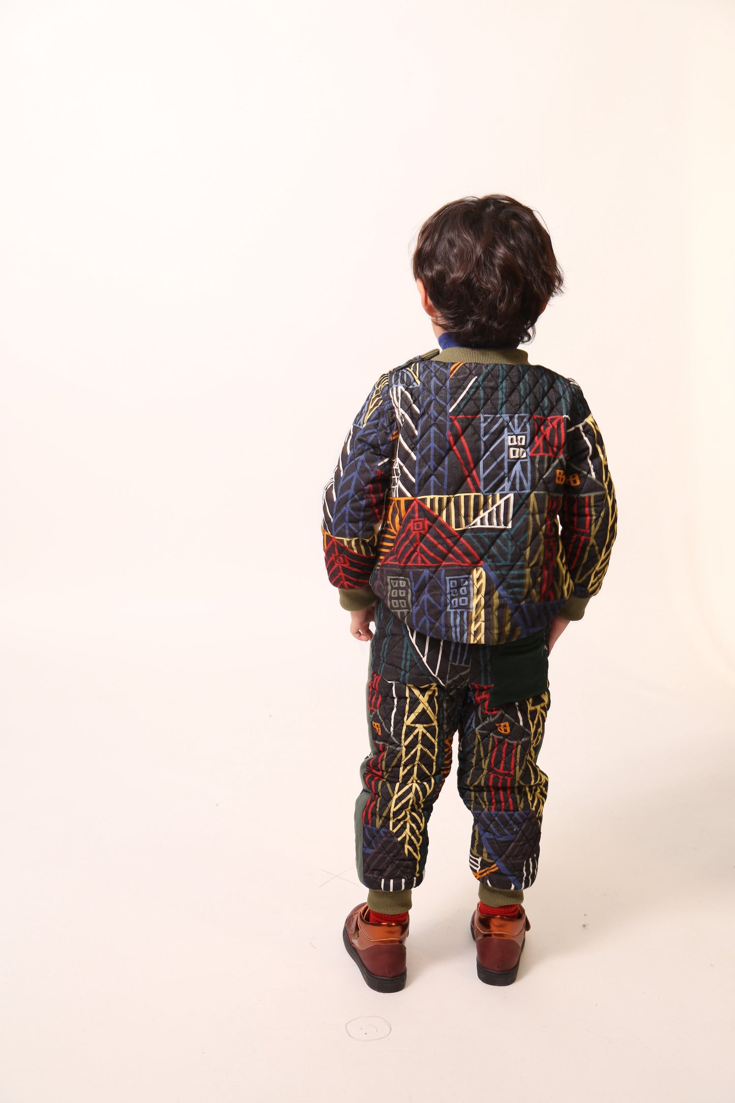 Boy wearing sporty track pants for boys in Normanz print, with harlequin diamond tufting and heavy padding for warmth. Pull-on elastic ribbed waistband, side seam pockets, ribbed leg openings for a snug fit. Contrast patch pocket at back, ribbons lining sides. Pair with Harlequin Padded Zip Sweatshirt for full rainbow track suit look. Dark rainbow, Tia Cibani.