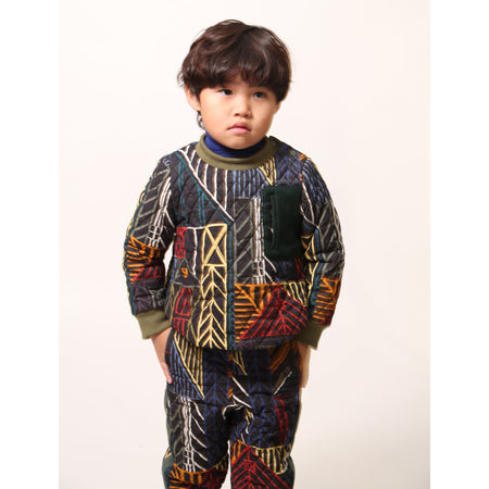 Boy wearing sporty track pants for boys in Normanz print, with harlequin diamond tufting and heavy padding for warmth. Pull-on elastic ribbed waistband, side seam pockets, ribbed leg openings for a snug fit. Contrast patch pocket at back, ribbons lining sides. Pair with Harlequin Padded Zip Sweatshirt for full rainbow track suit look. Dark rainbow, Tia Cibani.