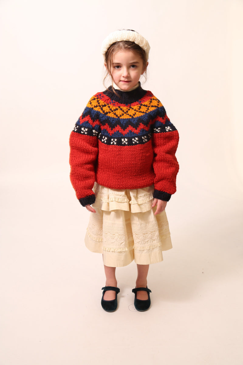 Girl wearing Long-sleeve sweater primarily red, with accents of blue and orange, made of Yarn fabric / Girl wearing a Tealength two-tiered taffeta skirt with elasticized smocking at the waistband, embroidered lace trim, and ribbon at each tier. Unlined for lightweight feel. Colour: Opal, by Tia Cibani. Front View.