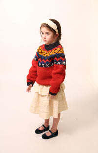 Girl wearing Long-sleeve sweater primarily red, with accents of blue and orange, made of Yarn fabric / Girl wearing a Tealength two-tiered taffeta skirt with elasticized smocking at the waistband, embroidered lace trim, and ribbon at each tier. Unlined for lightweight feel. Colour: Opal, by Tia Cibani. Front View.