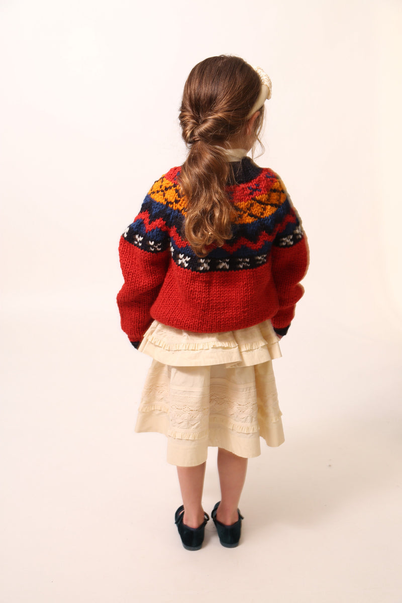 Girl wearing Long-sleeve sweater primarily red, with accents of blue and orange, made of Yarn fabric / Girl wearing a Tealength two-tiered taffeta skirt with elasticized smocking at the waistband, embroidered lace trim, and ribbon at each tier. Unlined for lightweight feel. Colour: Opal, by Tia Cibani. Front View.