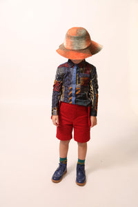 Boy wearing a collared shirt in Rainbow Normanz print, featuring colorful trims and blue snap tape detailing down the arms and at center front for button enclosure. Made from 100% Cotton, versatile for both casual and dressy settings, by Tia Cibani.