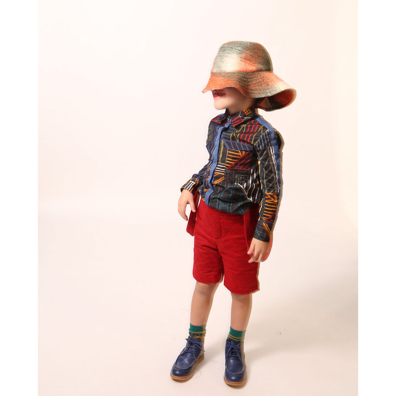 Boy wearing a collared shirt in Rainbow Normanz print, featuring colorful trims and blue snap tape detailing down the arms and at center front for button enclosure. Made from 100% Cotton, versatile for both casual and dressy settings, by Tia Cibani.