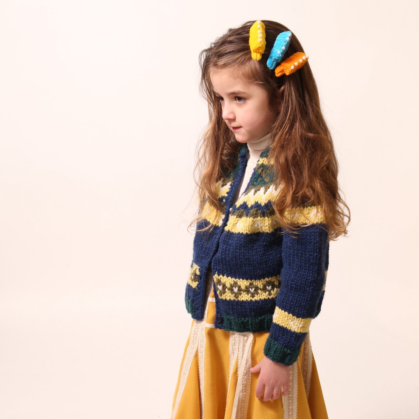 Girl wearing a long-sleeve cardigan in Yarn fabric, central button closure, predominantly blue with yellow and green accents, by Tia Cibani.