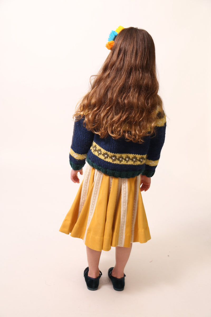 Girl photographed from behind, wearing a long-sleeve cardigan in Yarn fabric, central button closure, predominantly blue with yellow and green accents, by Tia Cibani.