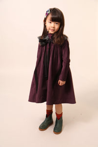 Girl wearing an oversized night shirt style dress, pin-tucked at the top with a darling high-low hem, featuring a detachable embroidered lace pleated collar at the mock neckline. Made from velvet fabric in Prune color, ideal for occasional wear or everyday dress up, with snap buttons and handy pockets at the sides, by Tia Cibani.