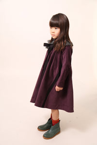 Girl wearing an oversized night shirt style dress, pin-tucked at the top with a darling high-low hem, featuring a detachable embroidered lace pleated collar at the mock neckline. Made from velvet fabric in Prune color, ideal for occasional wear or everyday dress up, with snap buttons and handy pockets at the sides, by Tia Cibani.