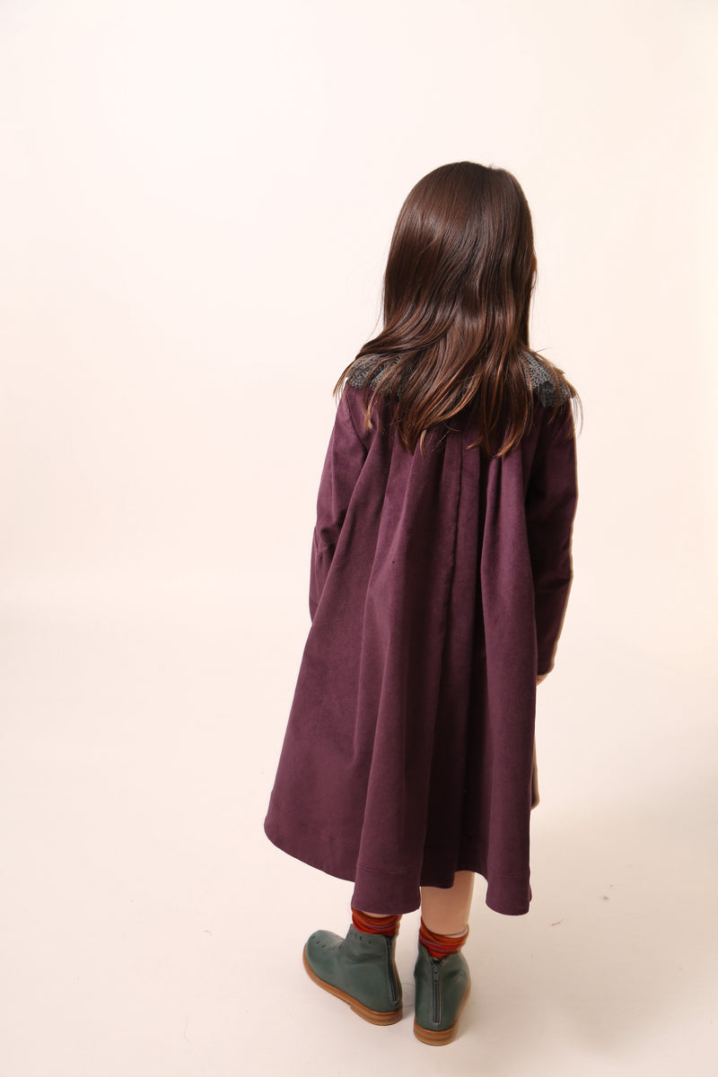 Girl wearing an oversized night shirt style dress, pin-tucked at the top with a darling high-low hem, featuring a detachable embroidered lace pleated collar at the mock neckline. Made from velvet fabric in Prune color, ideal for occasional wear or everyday dress up, with snap buttons and handy pockets at the sides, by Tia Cibani. Back View.