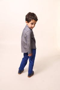 Boy wearing slim-fitting trousers in structured velvet, featuring subtle patchwork at hips and pocket flaps. Includes functional snaps, elastic waistband with adjustable button and loop, and zip-fly with hook and eye closure. Made from 99% Cotton, 1% Elastane with 100% Cotton lining. Color: Blue. / Boy wearing The Conan Embroidered Shirt, featuring high-low scoop hem and two-toned embroidered yoke. Collared with snap closure at center front. Perfect for casual and formal occasions. Colour: Blue,Tia Cibani.