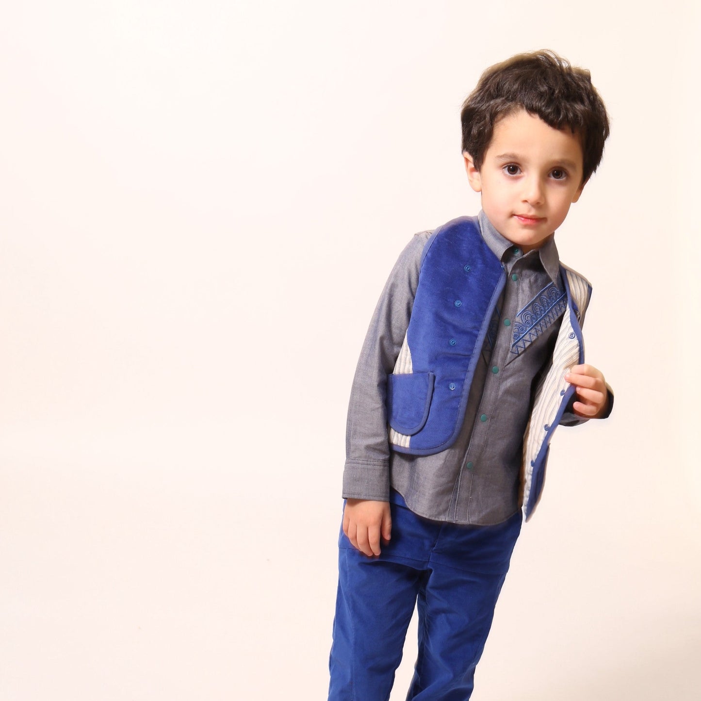 Boy wearing a versatile padded patchwork vest for boys, combining style and comfort with easy box fit for layering. Features binding tape at edges, patch pockets for treasures, and snap button closure at center front flap. Fully faced in cotton striped shirting, patchworked with structured velvet exterior for added fun. Great for formal and casual occasions. Pair with classic boy's button-ups and Breton Slim Fit Trousers for refined look. Blue and white, Tia Cibani.