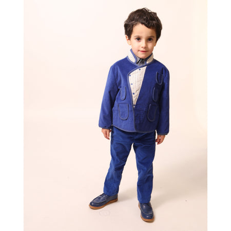 Boy wearing slim-fitting trousers in structured velvet, featuring subtle patchwork at hips and pocket flaps. Includes functional snaps, elastic waistband with adjustable button and loop, and zip-fly with hook and eye closure. Made from 99% Cotton, 1% Elastane with 100% Cotton lining. Color: Blue.