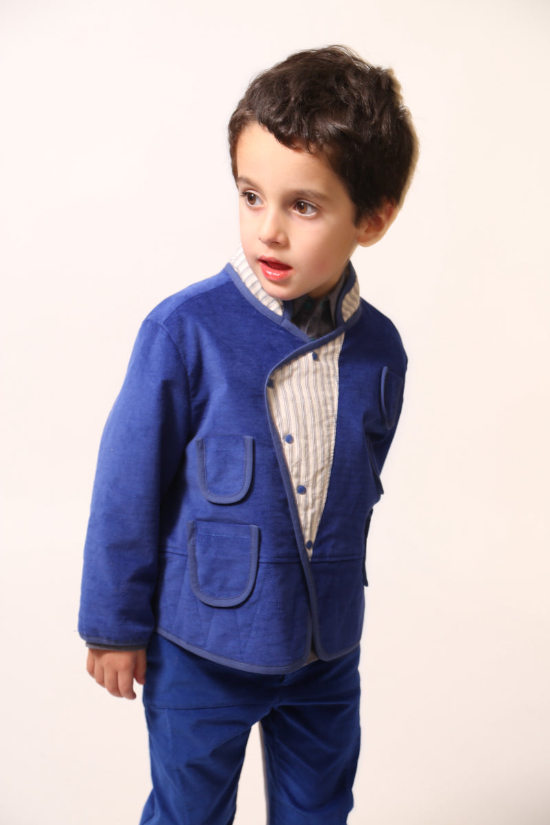 Boy wearing a padded patchwork jacket for boys with easy relaxed fit for comfort and layering. Double snap placket closure adds military-inspired flair. Fully faced with soft cotton striped shirting and patchworked with structured velvet on the outside. Features multiple patch pockets for treasures, binding tape trim throughout, and stand-up collar. Pair with Breton Slim Fit Trousers for refined look. Blue and white, Tia Cibani.