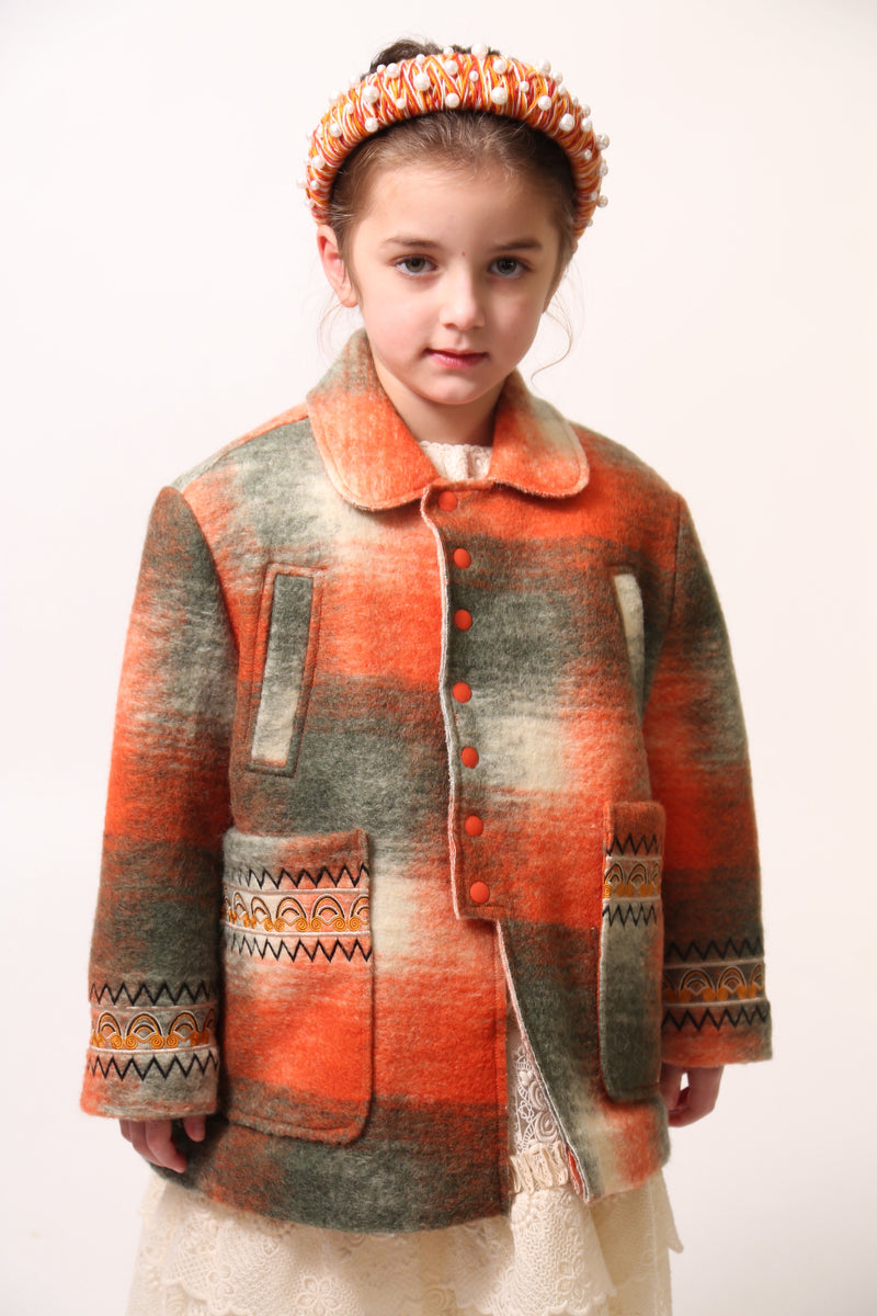 Girl wearing Loden style jacket in cozy Celtic Shadow Plaid, padded and lined for warmth. Snap closure, boxy yet tailored fit with sharp collar, roomy front pockets, and embroidery design at sleeve cuffs. Colour: Multicolour, mainly orange, Tia Cibani.