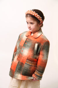 Girl wearing Loden style jacket in cozy Celtic Shadow Plaid, padded and lined for warmth. Snap closure, boxy yet tailored fit with sharp collar, roomy front pockets, and embroidery design at sleeve cuffs. Colour: Multicolour, mainly orange, Tia Cibani.