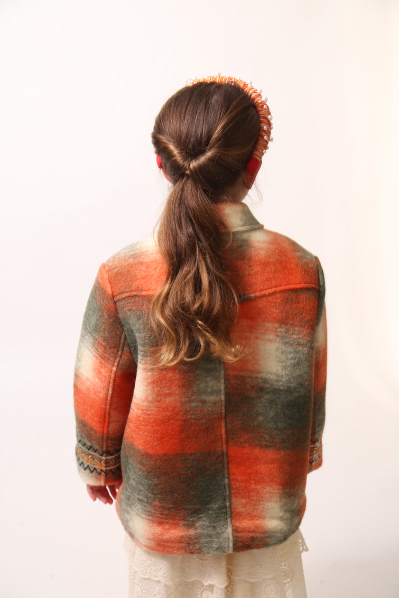 Girl wearing Loden style jacket in cozy Celtic Shadow Plaid, padded and lined for warmth. Snap closure, boxy yet tailored fit with sharp collar, roomy front pockets, and embroidery design at sleeve cuffs. Colour: Multicolour, mainly orange, Tia Cibani. Back View.