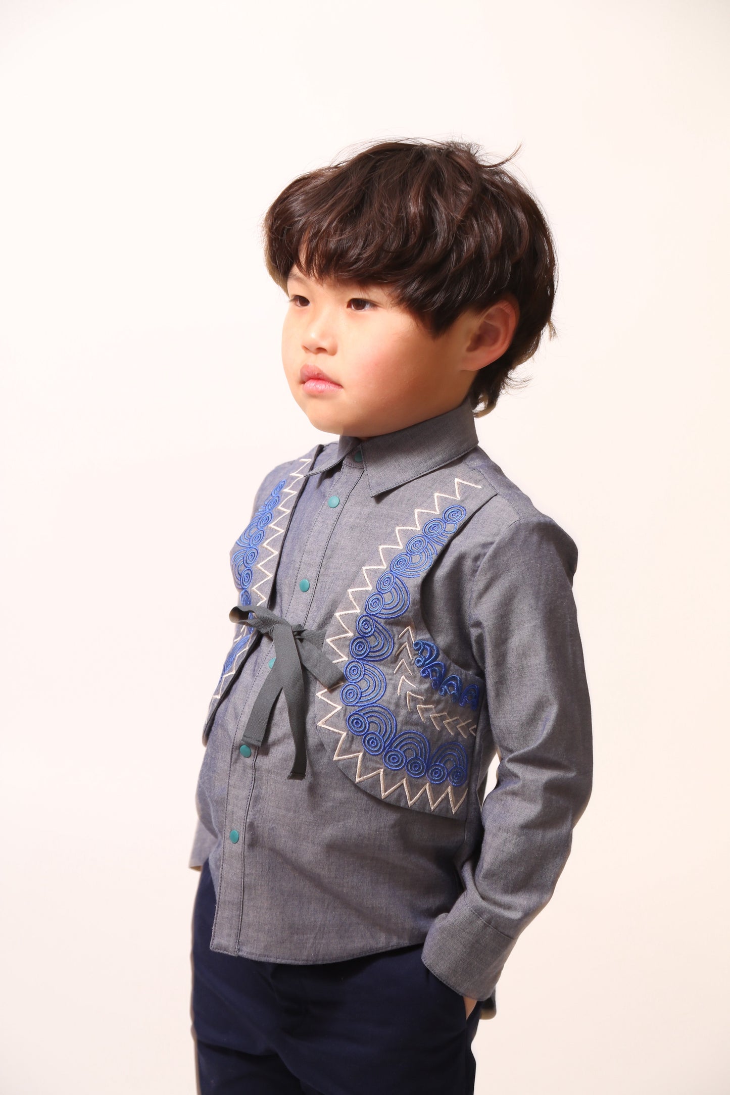 Boy wearing a western-inspired take on classic boy's button-up with scoop high-low hem for modern twist. Built-in vest with two-toned embroidery design and textured ribbons at overlay. Snap closure at center front for easy dressing. Perfect for any occasion, from family gatherings to formal events. Cotton. Blue, Tia Cibani. 