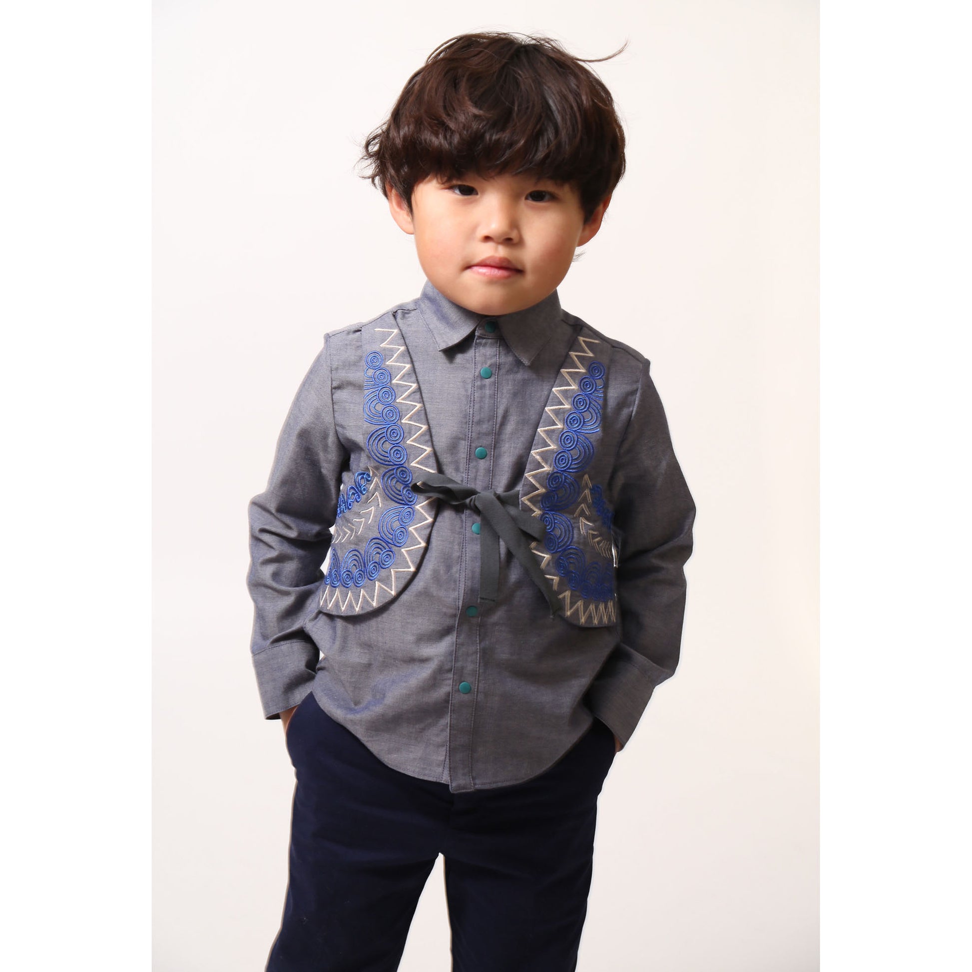 Boy wearing a western-inspired take on classic boy's button-up with scoop high-low hem for modern twist. Built-in vest with two-toned embroidery design and textured ribbons at overlay. Snap closure at center front for easy dressing. Perfect for any occasion, from family gatherings to formal events. Cotton. Blue, Tia Cibani. 