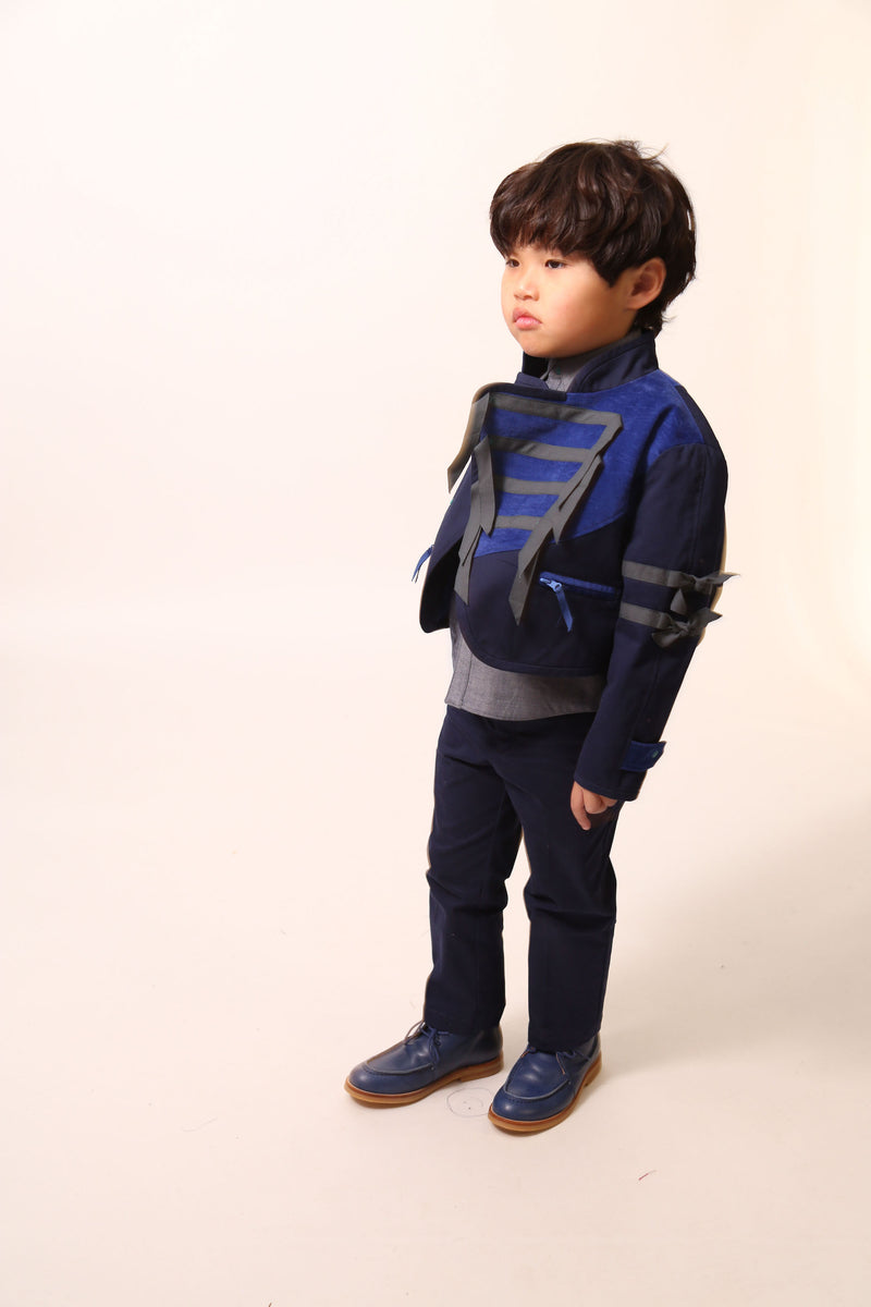 Bold and stylish Malok Military Jacket for boys, inspired by traditional military dress. Patchworked with cotton twill and velvet accents. Features zip welt pockets, yoke at back, snap tabs at sleeves, and double-breasted snap closure for comfort and charm. Relaxed box fit for easy layering. Textured structured ribbon details on front and sleeves with free-hanging tassels for contemporary flair. Scoop hem, stand-up collar. Perfect with Malok Slim Fit Trousers. Blue, Tia Cibani.