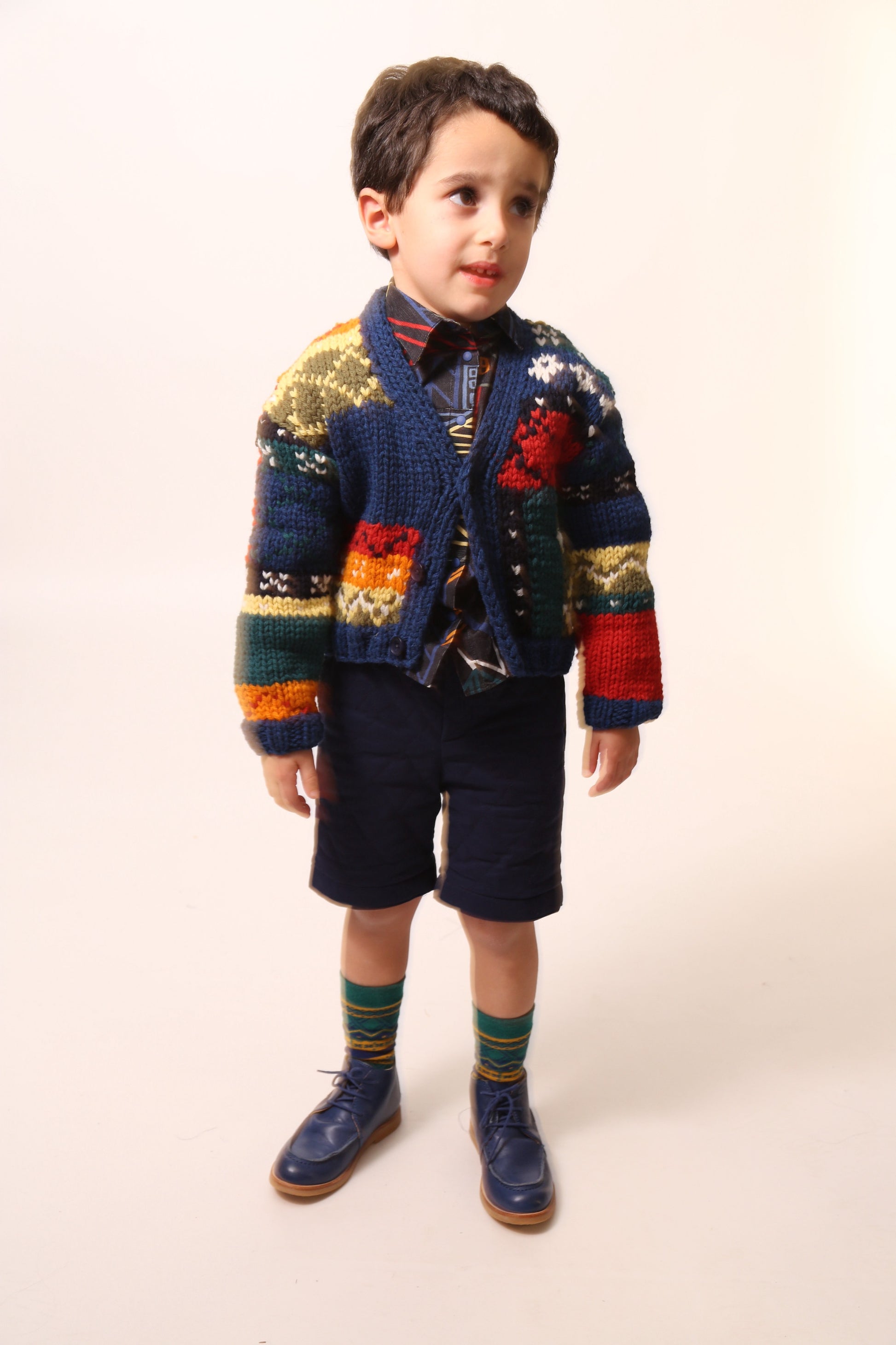 Boy wearing  a colorful fair isle cardigan in a patchwork design, blending various fair isle patterns with solid colors of Felt Wool Yarn. Features a relaxed fit with faux horn buttons at the center front, ideal for layering over basic knits. Made from 100% Wool. Color: Multicolor. By Tia Cibani.