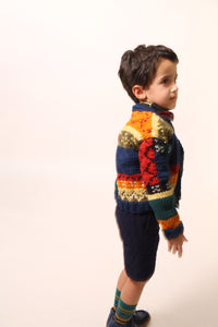 Boy wearing  a colorful fair isle cardigan in a patchwork design, blending various fair isle patterns with solid colors of Felt Wool Yarn. Features a relaxed fit with faux horn buttons at the center front, ideal for layering over basic knits. Made from 100% Wool. Color: Multicolor. By Tia Cibani.