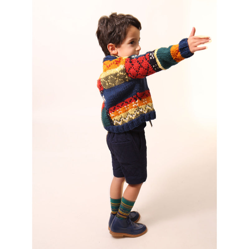 Boy wearing  a colorful fair isle cardigan in a patchwork design, blending various fair isle patterns with solid colors of Felt Wool Yarn. Features a relaxed fit with faux horn buttons at the center front, ideal for layering over basic knits. Made from 100% Wool. Color: Multicolor. By Tia Cibani.