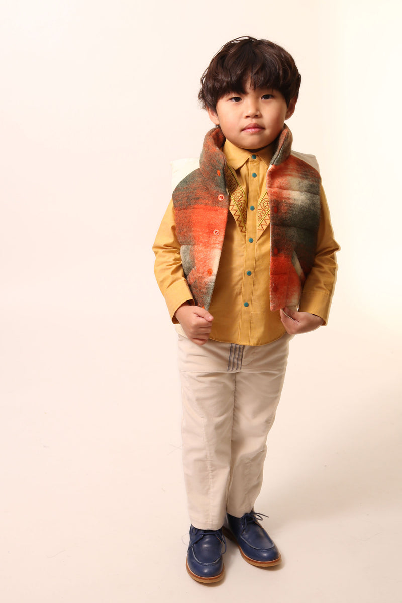 Boy wearing a sleeveless vest. Contrast color snaps at center front add pop of color and flair. Slanted pockets with pocket bag peering through at hem for style and warmth. Hint of taffeta at shoulders and back hemline for patchwork effect. Down-filled with high collar for warmth. Ideal for layering basic knits or thicker sweaters. Wool. Orange,Tia Cibani.