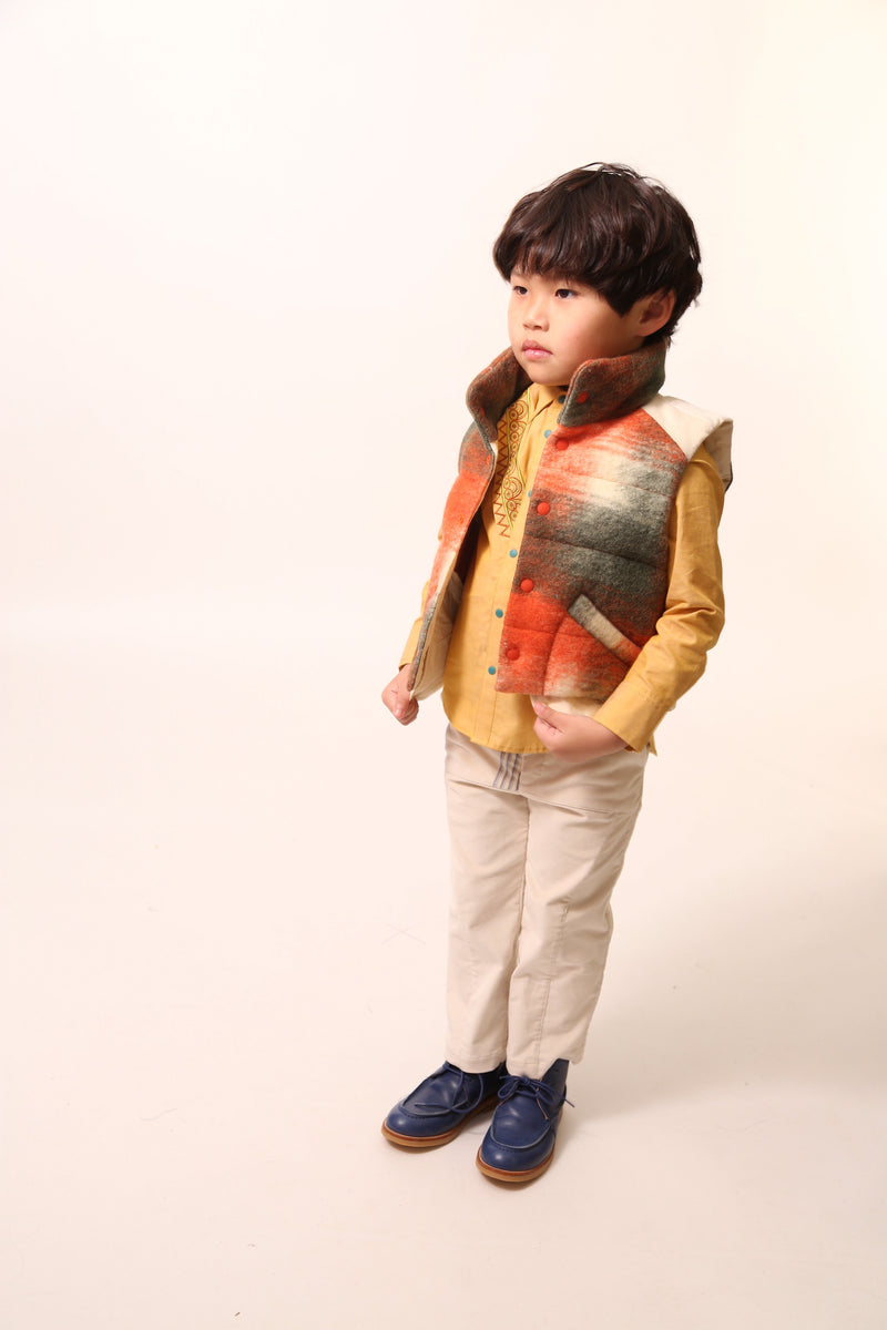 Boy wearing a sleeveless vest. Contrast color snaps at center front add pop of color and flair. Slanted pockets with pocket bag peering through at hem for style and warmth. Hint of taffeta at shoulders and back hemline for patchwork effect. Down-filled with high collar for warmth. Ideal for layering basic knits or thicker sweaters. Wool. Orange,Tia Cibani.