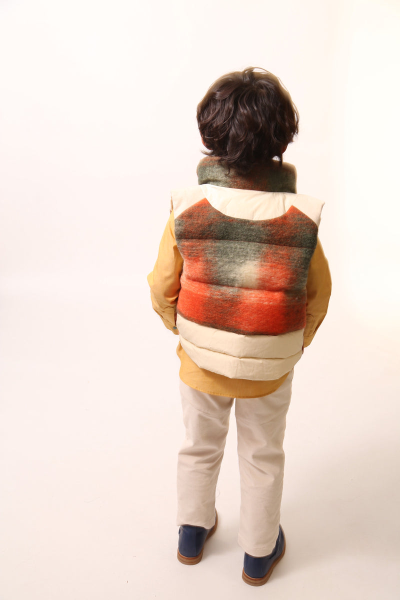 Boy wearing a sleeveless vest. Contrast color snaps at center front add pop of color and flair. Slanted pockets with pocket bag peering through at hem for style and warmth. Hint of taffeta at shoulders and back hemline for patchwork effect. Down-filled with high collar for warmth. Ideal for layering basic knits or thicker sweaters. Wool. Orange,Tia Cibani.