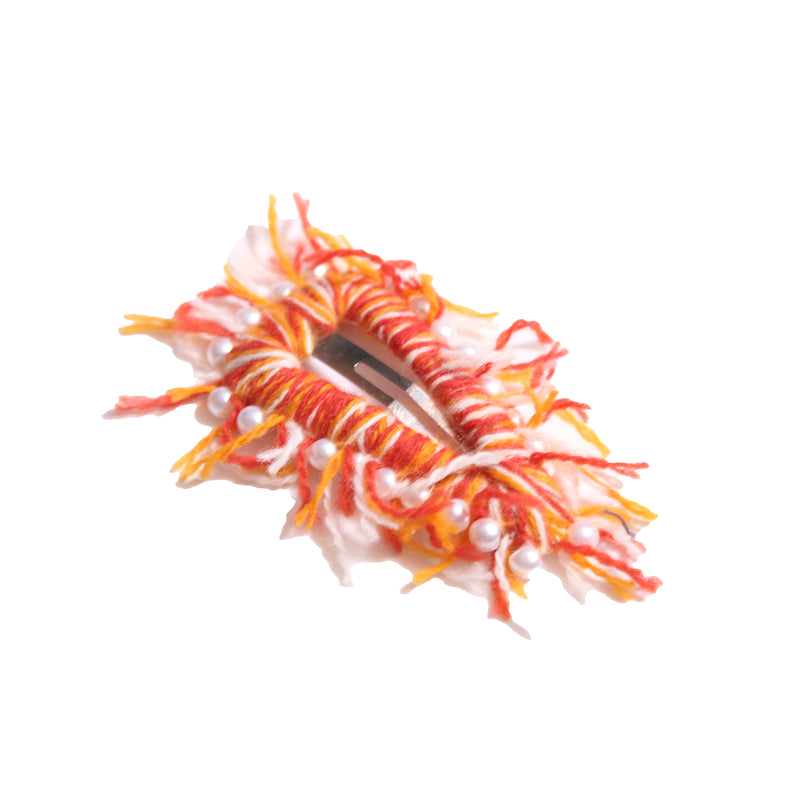 Oversized hair clip with a sturdy metal snap clip base, wrapped and fringed in colorful red and orange wool yarn, decorated with pearls for a fun finish, by Tia Cibani.